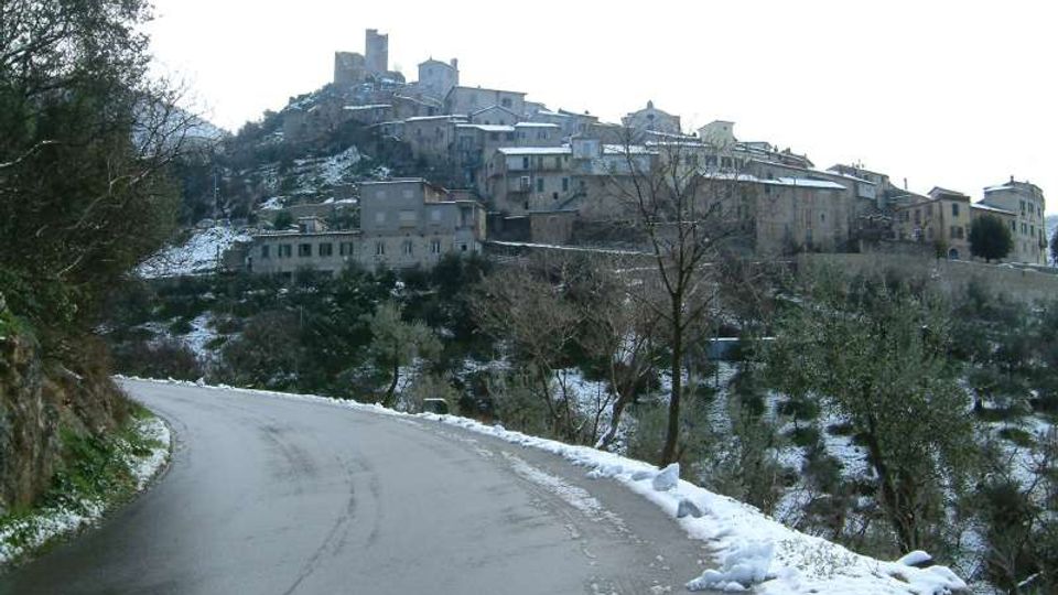 Strada asfaltata in curva con bordi innevati che conduce verso un borgo medievale arroccato su una collina, con case in pietra e torri in cima. Vegetazione di alberi e pendii con chiazze di neve circondano l’abitato.