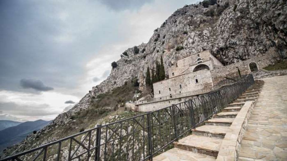 Scalinata in pietra con ringhiera metallica che sale lungo un pendio roccioso verso un edificio storico in pietra addossato alla montagna. Sullo sfondo si vedono pareti di roccia scoscesa, vegetazione rada e cielo nuvoloso.