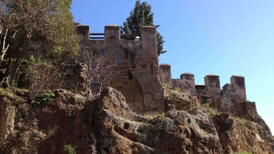 Ruderi di mura fortificate in pietra con merli e torrette, arroccati su una scogliera rocciosa con vegetazione spontanea. Sullo sfondo cielo sereno e alcuni alberi dietro la struttura.