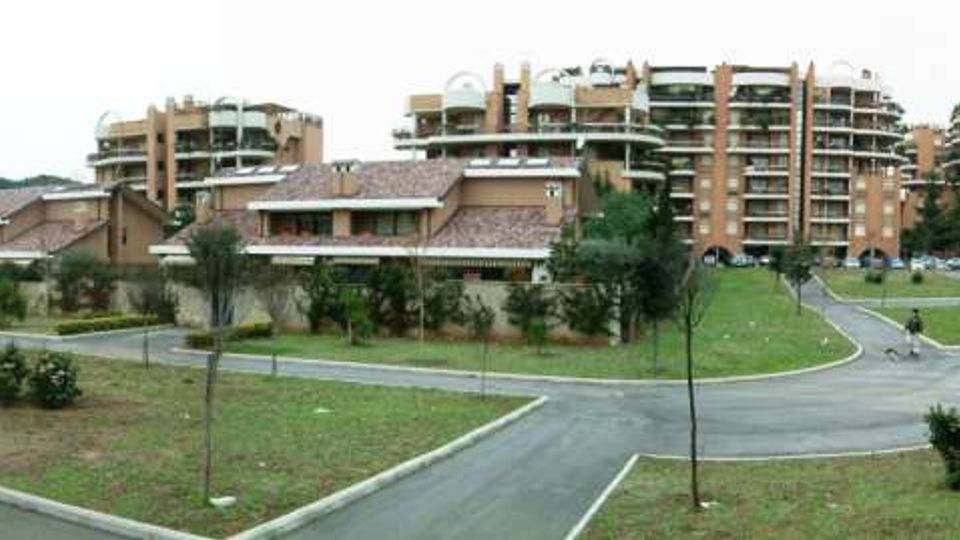 Panorama di un complesso residenziale con alti condomini in mattoni e ampi balconi, affiancati da edifici più bassi con tetti a falde. In primo piano si vedono strade interne asfaltate e aree verdi con prati e alberi giovani, disposte tra i percorsi.