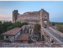 Castello in pietra su uno sperone roccioso, con mura alte e torri rettangolari, collegato da un ponte in muratura che conduce a un portale d’ingresso ad arco. In primo piano si vedono tetti in coppi di edifici sottostanti e vegetazione circostante, con cielo sereno sullo sfondo.