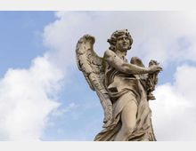 Statua in pietra di un angelo alato in stile classico, con drappeggi e braccia tese, ripresa dal basso contro un cielo azzurro con nuvole.