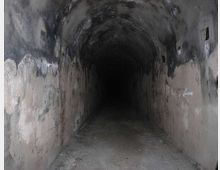 Interno di un lungo tunnel sotterraneo con volta a botte, pareti in cemento macchiate e annerite e pavimento polveroso. Piccole aperture laterali interrompono le pareti mentre il corridoio si perde nel buio in fondo.