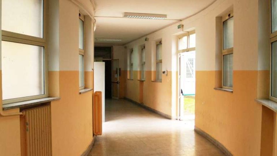 Corridoio interno curvo di un edificio pubblico, con pareti bicolori crema e arancione, pavimento in piastrelle grigie e finestre rettangolari lungo entrambi i lati. Illuminazione a plafoniere sul soffitto e una porta aperta in fondo che lascia entrare luce dall’esterno.