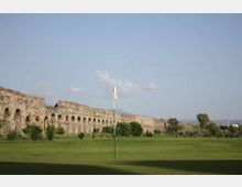 Campo da golf con ampia distesa erbosa e bandierina su asta al centro; sullo sfondo si estende una lunga struttura in pietra con arcate, simile a un acquedotto o muro storico, con alberi sparsi e cielo sereno.