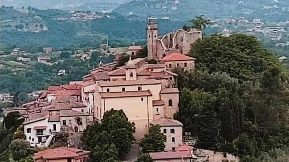 Veduta panoramica di un borgo collinare con case dai tetti in coppi rossi e una strada centrale alberata che sale verso una chiesa con campanile. Sullo sfondo si vedono colline e montagne verdi con abitazioni sparse nella valle.