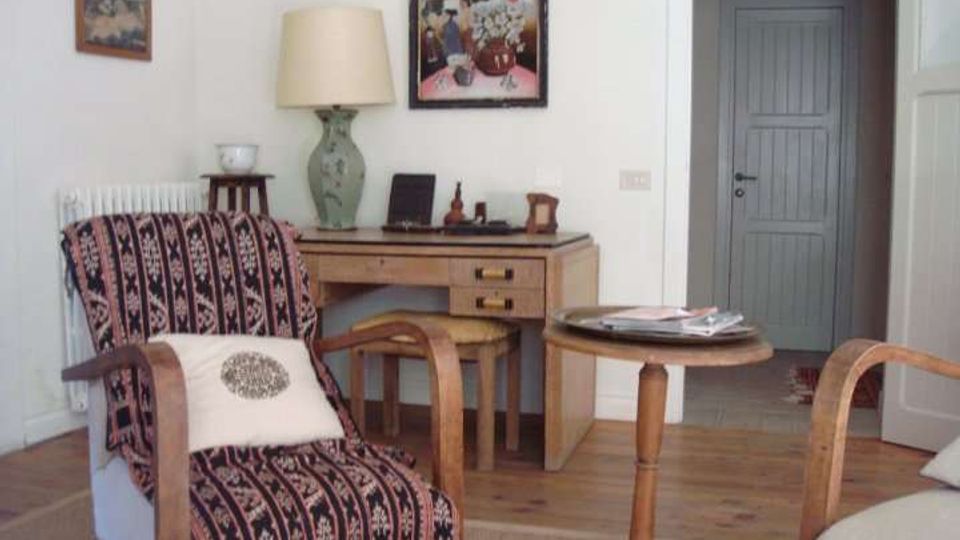 Soggiorno arredato con poltrona in legno rivestita in tessuto a motivi, piccolo tavolino rotondo e scrivania con lampada e decorazioni, con quadri appesi alla parete. Sullo sfondo si vede un’apertura che conduce a un corridoio con porta chiusa.