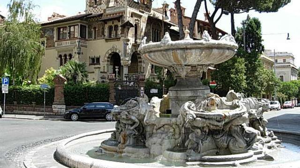 Fontana monumentale in una rotonda stradale, con grande vasca circolare e gruppo scultoreo in pietra attorno alla base. Sullo sfondo si vede una villa storica in stile eclettico con torre e logge, circondata da alberi alti e vegetazione urbana.