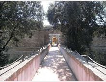 Passerella pedonale in muratura con corrimano in corda che conduce a un arco d’ingresso in pietra su un alto muro fortificato. Ai lati si vedono alberi e vegetazione che incorniciano il percorso.