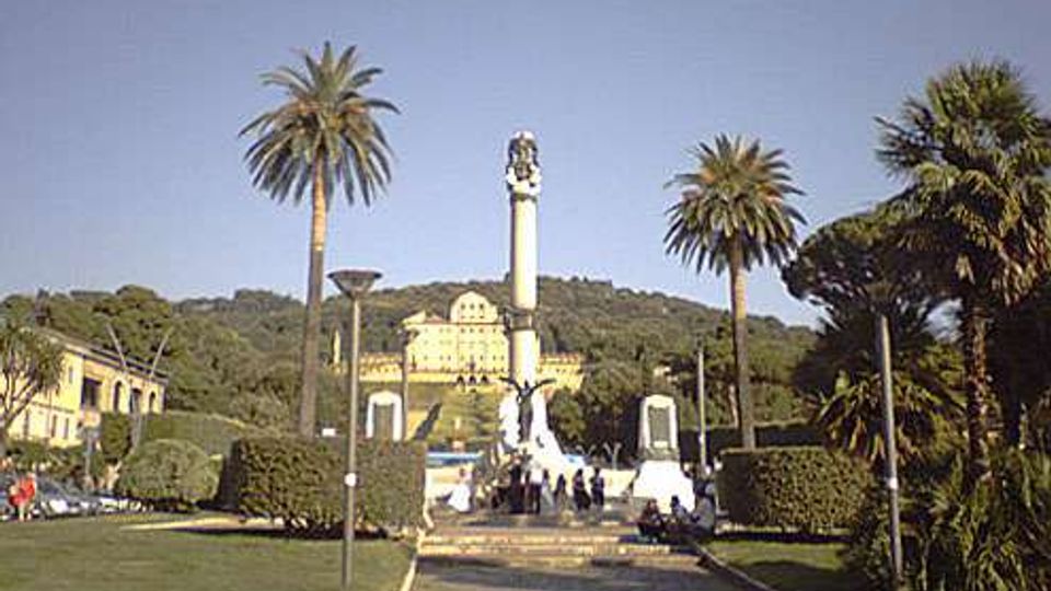 Piazza con ampio prato e scalinata centrale che conduce a un alto monumento su piedistallo, circondato da palme e siepi potate. Sullo sfondo si vedono edifici chiari e una collina boscosa sotto un cielo sereno.