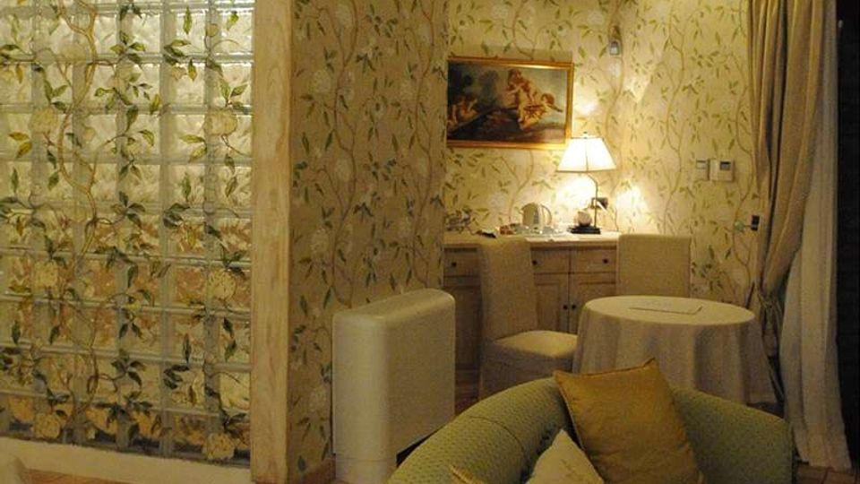 Interno di una camera o suite con arredi classici: pareti con carta da parati a motivi vegetali, pavimento in piastrelle e soffitto con travi a vista. Un divisorio in mattoni di vetro separa l’area soggiorno con poltrona imbottita, tavolino rotondo, credenza con lampada e grandi tende davanti a una porta-finestra.