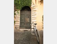 Ingresso di un edificio con grande portone in legno scuro sotto un arco in pietra bugnata e grata semicircolare in ferro, con edera che ricopre parte della facciata. Davanti al portone, su pavimentazione in sampietrini, è appoggiata una bicicletta.