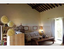 Soggiorno in stile rustico con soffitto alto e travi in legno a vista, divano con cuscini decorati e grande tavolino basso in legno. A sinistra si vede un angolo cucina con scaffali e bancone, mentre a destra una porta-finestra con tende bianche apre su un balcone.