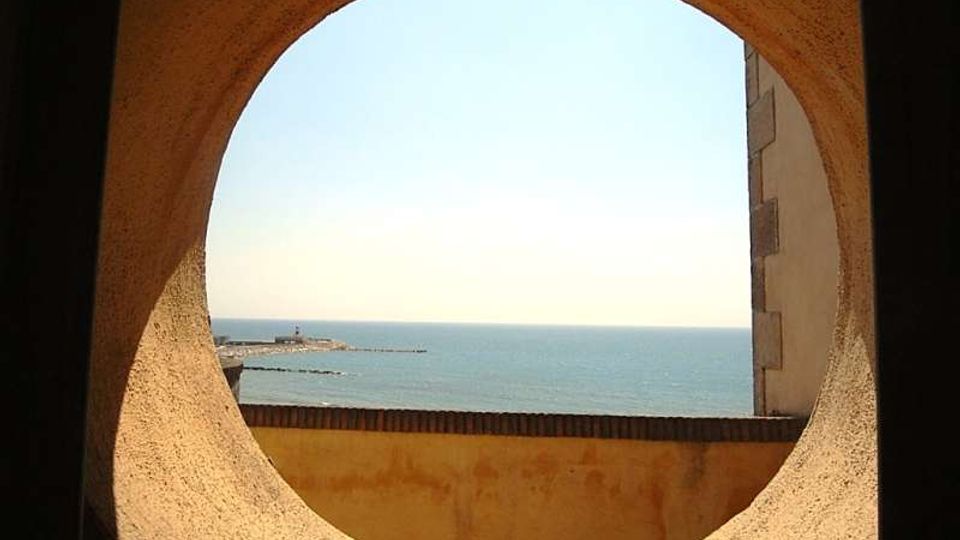 Veduta del mare attraverso un grande foro circolare in una parete in muratura, con bordi intonacati e un muretto in primo piano. All’orizzonte si vede una diga o molo con un piccolo faro e il cielo limpido sullo sfondo.