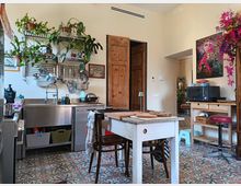 Cucina abitata con pavimento in piastrelle decorative e isola centrale in legno bianco con piano in marmo e sgabelli. A sinistra ci sono lavello e piano in acciaio con mensole piene di piante e utensili appesi, mentre a destra un carrello con microonde e una porta in legno conduce a un altro ambiente.