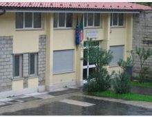 Edificio pubblico a due piani con facciata giallo chiaro e parti in muratura grezza, tetto in tegole rosse e finestre con serrande metalliche. All’ingresso con porta vetrata sono presenti bandiere e un cartello, davanti a un piccolo giardino con arbusti e un piazzale asfaltato bagnato.