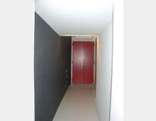 Corridoio interno stretto con pavimento chiaro, parete sinistra scura e parete destra bianca, che conduce a una porta doppia rossa in fondo. Soffitto basso con piccola griglia o vano di aerazione sopra la porta.