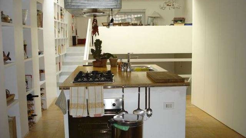 Cucina moderna in ambiente open space con isola centrale in legno dotata di piano cottura, lavello e cappa in metallo sospesa. Ai lati si vedono scaffalature bianche a giorno e una parete divisoria in vetro satinato, con pavimento in legno chiaro e arredi minimalisti.