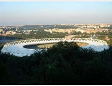 Vista panoramica di uno stadio ovale con struttura esterna bianca a traliccio, circondato da alberi in primo piano. Sullo sfondo si vede un’ampia area urbana con edifici e zone verdi sotto un cielo sereno.