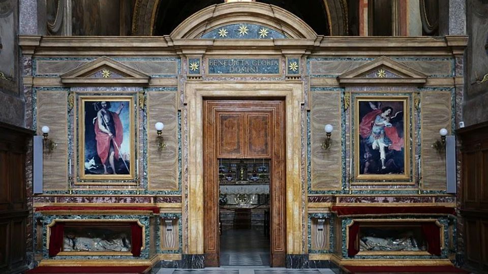 Interno di una chiesa con parete in marmi policromi e portale centrale ad arco che conduce a una cappella. Ai lati del portale ci sono due grandi dipinti incorniciati, lampade a parete e nicchie con sarcofagi, sopra un’iscrizione decorata con stelle e una piccola finestra circolare.
