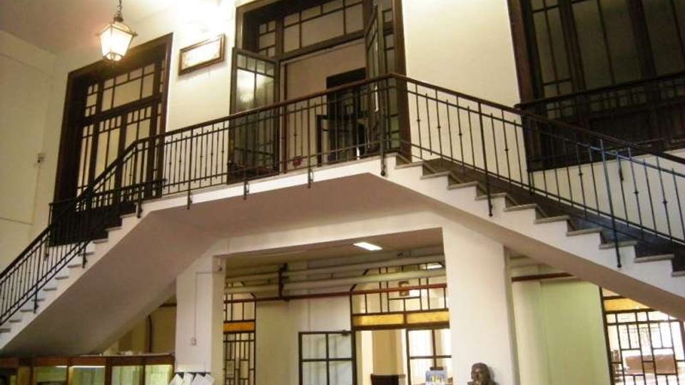 Interno di un edificio con scalinate simmetriche che salgono a un ballatoio centrale con ringhiere in ferro battuto. Grandi finestre e porte in legno e vetro con riquadri geometrici, illuminazione a lampada sospesa e colonne bianche al piano terra con vetrine e una statua su piedistallo.