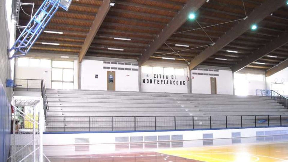 Interno di un palazzetto dello sport con campo da basket in parquet e canestri, affacciato su grandi gradinate in cemento con corrimano. Il soffitto è alto con travi in legno e luci sospese, e sulla parete di fondo compare la scritta “Città di Montefiascone”.
