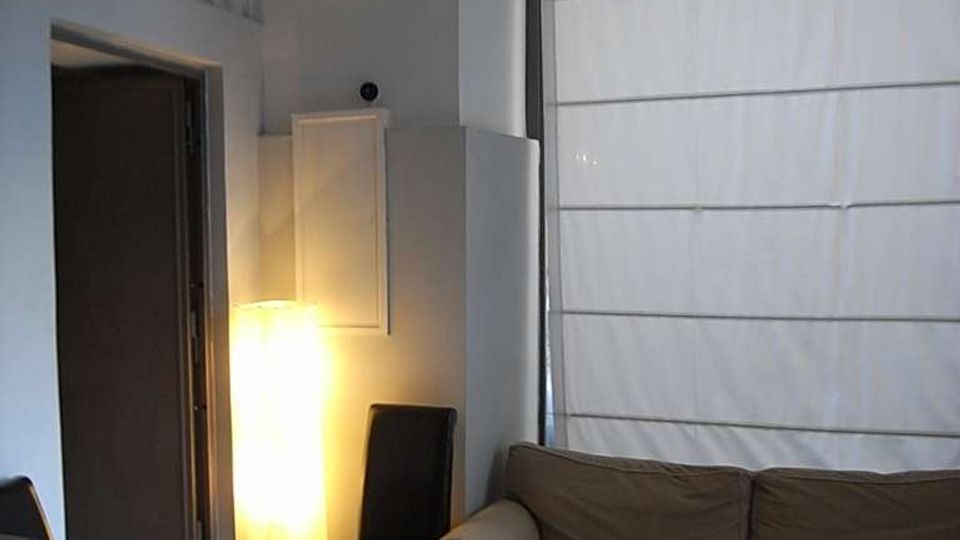 Interno di un piccolo soggiorno con divano grigio, sedie nere e lampada da terra accesa su pavimento in graniglia. Una grande finestra ad arco con inferriata decorativa e tenda a pannelli occupa la parete destra, accanto a una porta aperta su un corridoio.