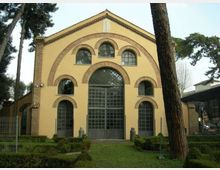 Facciata di un edificio in stile industriale con intonaco giallo e archi in mattoni, con grande portale centrale vetrato e finestre ad arco simmetriche. Davanti si vede un prato con siepi basse e alberi alti che incorniciano l’ingresso.