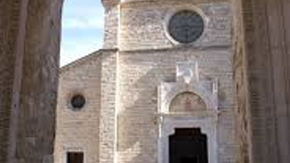 Vista attraverso un arco in pietra di una chiesa in muratura chiara con facciata semplice, rosone circolare e portale centrale con timpano. Davanti all’ingresso si apre un piccolo sagrato lastricato illuminato dal sole.