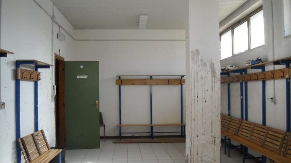 Spogliatoio semplice con pavimento piastrellato e soffitto bianco, dotato di panche in legno, appendiabiti e scaffali lungo le pareti. Una colonna centrale divide lo spazio; sul lato destro ci sono finestre alte che lasciano entrare luce naturale e una porta verde sul fondo.
