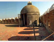 Terrazza in mattoni su una fortificazione con parapetti merlati e un piccolo edificio a pianta circolare con cupola in pietra e porta aperta. Lo spazio è all’aperto, affiancato da muri in mattoni e con vista sul cielo limpido.