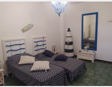 Camera da letto in stile marinaro con due letti singoli affiancati, testiere bianche con dettagli blu e biancheria scura a motivi. Sulla parete ci sono uno specchio con cornice blu e un mobile basso bianco, mentre nell’angolo si vede una decorazione a forma di faro; pavimento in piastrelle verdi e lampadario a soffitto.