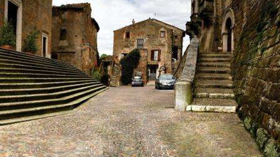Piazza in acciottolato in un borgo storico, circondata da edifici in pietra e mattoni con due ampie scalinate laterali. Sullo sfondo si vede una casa centrale e alcune auto parcheggiate sotto un cielo nuvoloso.
