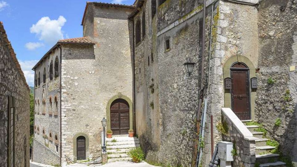Vicolo in pietra in un borgo storico, con edifici in muratura grezza e facciate alte affacciate su una piccola piazzetta. Si vedono porte ad arco, finestre strette, una scalinata con corrimano in pietra, lampioni a parete e pavimentazione in lastre sotto un cielo azzurro.
