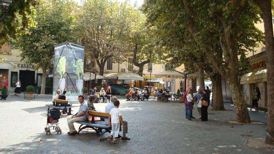 Piazza urbana lastricata con ampi alberi che creano ombra, panchine e una struttura a forma di chiosco vetrato al centro. Sullo sfondo si vedono edifici bassi con portici e ombrelloni di bar e ristoranti all’aperto.