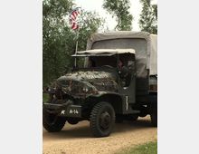 Camion militare d’epoca con telone sul cassone e rete mimetica sul cofano, fermo su una strada sterrata circondata da alberi. Una bandiera statunitense è montata su un’asta nella parte anteriore del veicolo.