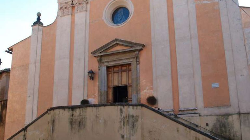 Facciata di una chiesa in stile classico con timpano triangolare e croce in cima, grandi paraste verticali e rosone circolare sopra il portale. Una scalinata con muretto conduce all’ingresso, con vasi di piante e strada lastricata in primo piano.