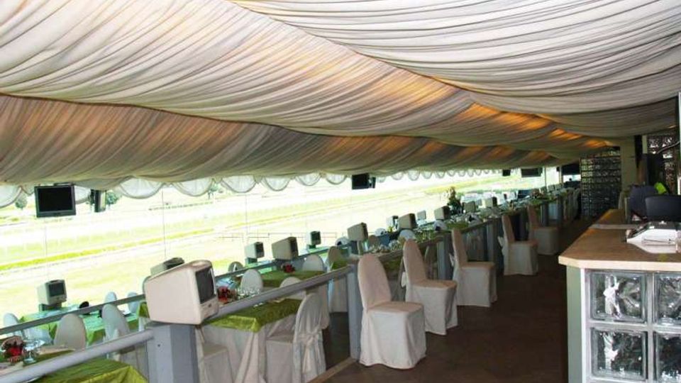 Area lounge coperta con grande soffitto in tessuto drappeggiato, tavoli apparecchiati con sedie rivestite e bancone con parete in vetrocemento. Lungo il perimetro ci sono vetrate affacciate su una pista all’aperto.