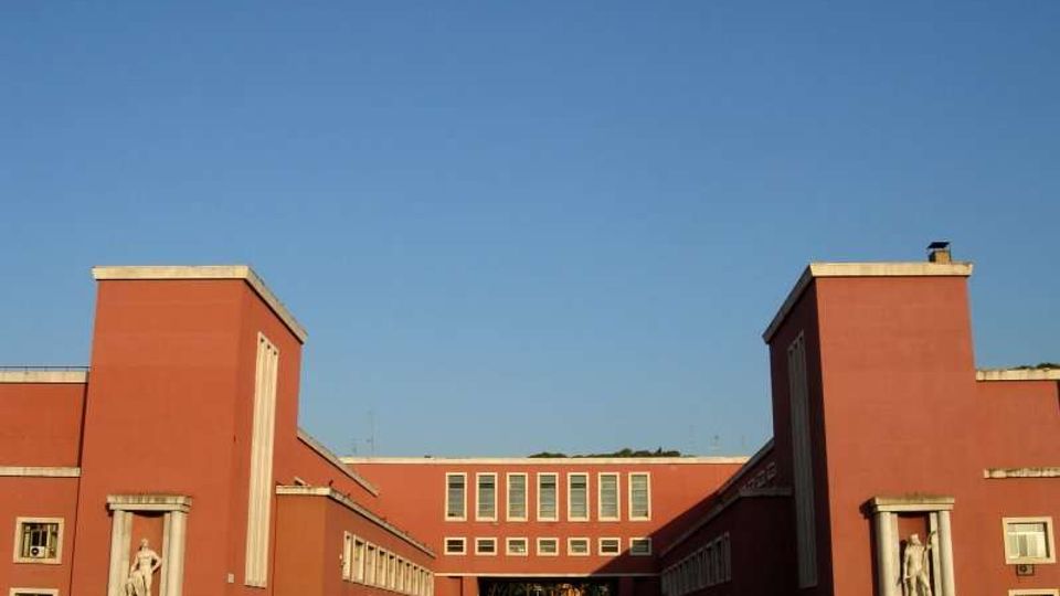 Edificio istituzionale in mattoni rossi con ali simmetriche e due torri laterali, caratterizzato da finestre rettangolari in file regolari. All’ingresso, nicchie con statue e un passaggio centrale verso il cortile interno sotto un cielo sereno.