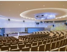 Ampio auditorium moderno con file di poltrone in legno disposte a semicerchio su gradinate, rivolte verso un palco con banco relatori. Soffitto curvo con illuminazione incassata e grande elemento luminoso ovale centrale, pareti bianche con apparecchi audio e luci.