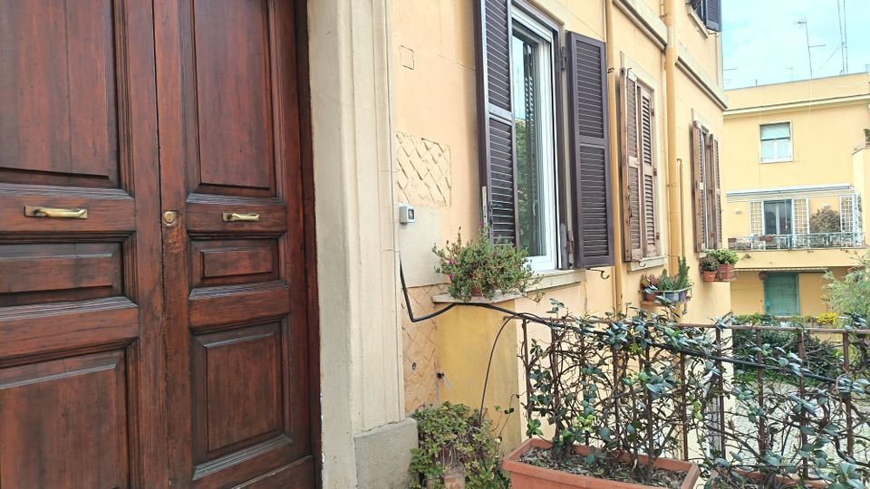 Ingresso di un edificio residenziale con grande portone in legno scuro e facciata color crema, affiancato da finestre con persiane scure e fioriere. In primo piano un piccolo balcone o pianerottolo con ringhiera metallica e vasi rettangolari di piante, con altri palazzi sullo sfondo.