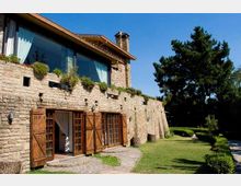 Casa in pietra a due piani con grandi finestre e portefinestre con persiane in legno, balconata con vasi di piante e tetto spiovente con camino. Davanti c’è un giardino curato con prato, siepi e vialetto in pietra, con alti alberi sullo sfondo.