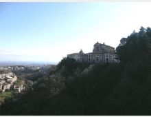 Veduta panoramica di un complesso religioso con chiesa e edifici adiacenti su un colle, affacciato su una valle. In primo piano si vedono pendii alberati, mentre a sinistra si estende un piccolo centro abitato con case in pietra.