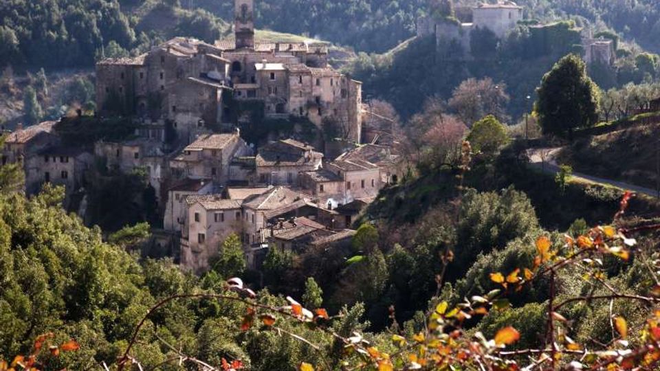 Veduta panoramica di un borgo storico in pietra su un pendio, con case addossate e un campanile centrale. Il villaggio è circondato da colline boscose, vegetazione in primo piano e un edificio fortificato su un’altura sullo sfondo.