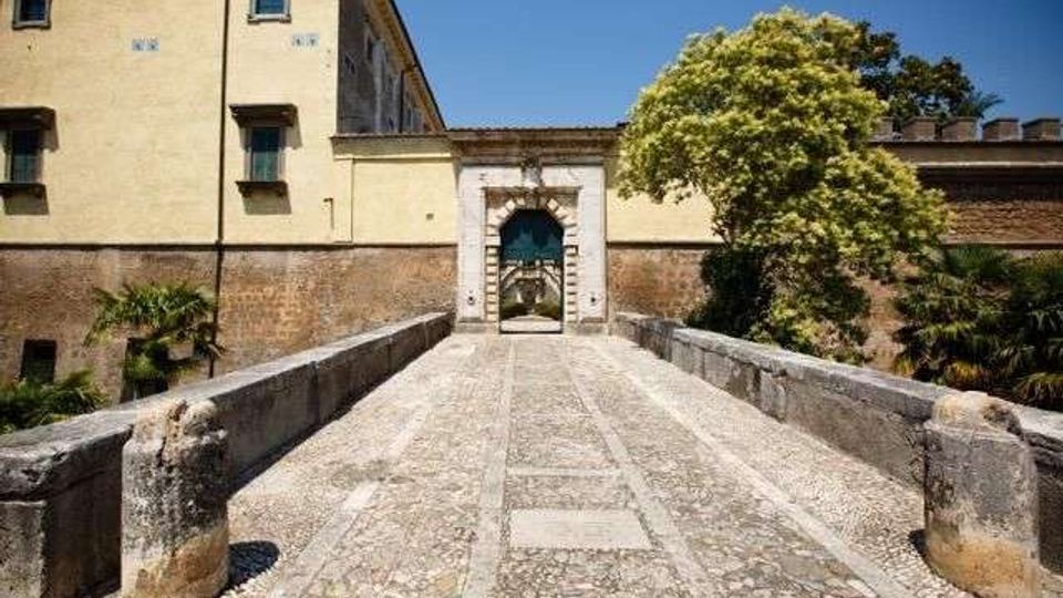 Ponte in pietra con pavimentazione a mosaico che conduce a un portale ad arco in muratura, inserito nelle mura di un edificio storico. Ai lati si vedono muri in mattoni e alberi, con cielo sereno sullo sfondo.