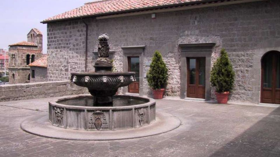 Cortile in pietra con fontana ottagonale in stile antico al centro, su pavimentazione lastricata. Sullo sfondo un edificio in muratura di pietra con tetto in coppi e porte in legno, affiancate da due vasi con arbusti.