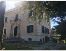 Villa storica a due piani con facciata chiara, persiane scure e ingresso con scalinata e portico a colonne. Intorno si vede un giardino con vialetto in pietra e alberi che incorniciano l’edificio, con una balaustra sul lato destro.