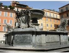 Fontana monumentale in pietra con grande vasca circolare centrale, base rettangolare e sculture decorative ai lati, collocata in una piazza urbana. Sullo sfondo si vedono palazzi storici a più piani con facciate colorate e finestre regolari.