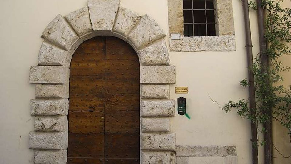 Facciata di un edificio con grande portone ad arco in legno, incorniciato da blocchi di pietra rustica. Accanto al portone c’è una piccola finestra quadrata con inferriata, con tubo di scarico e vegetazione rampicante sul lato destro.