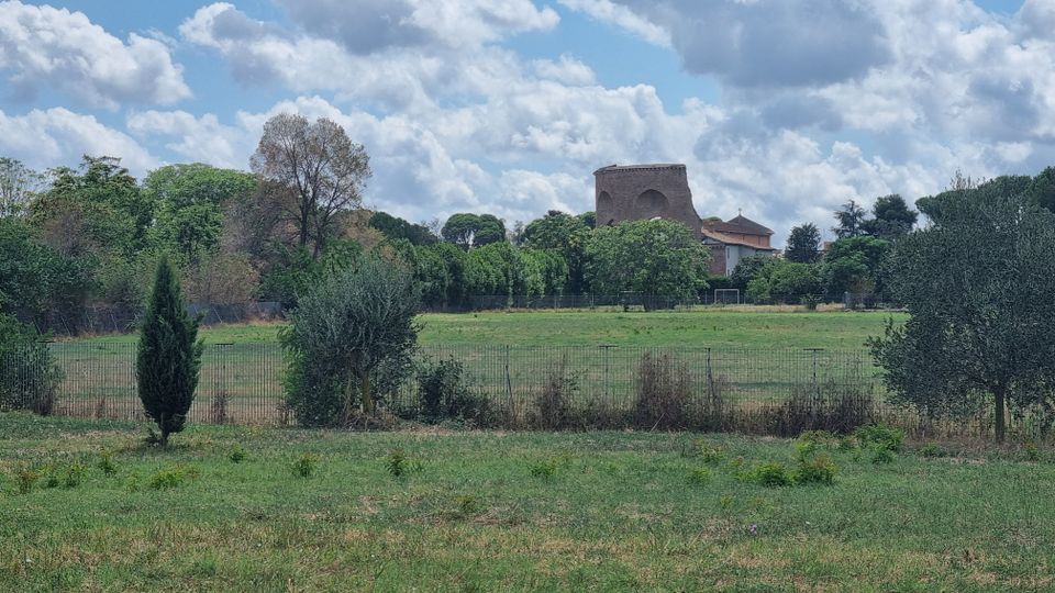 Ampio prato recintato con alberi e cespugli, sullo sfondo un complesso di edifici in muratura con una struttura alta e squadrata tra la vegetazione. Cielo azzurro con grandi nuvole sparse sopra un paesaggio rurale pianeggiante.